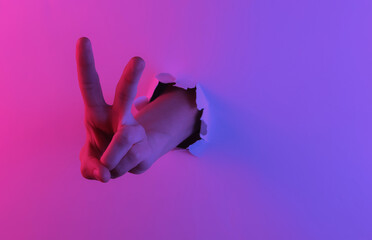 Mystical halloween concept. Hand shows V gesture through a torn hole..Creative pop art pink blue neon color. Trendy gradient illumination. Night light