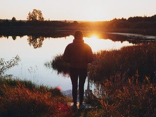 A girl in a warm jacket stands on the shore of the lake and looks into the distance at the sunset or sunrise. Silhouette of a girl in the bright sun. Beautiful romantic autumn landscape