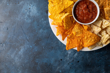 Wall Mural - Round white plate with corn tortilla chips and salsa on dark blue background. Unhealthy food. Concept of spicy meal. Top view.