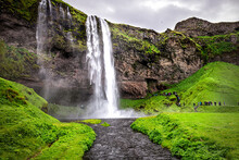Seljalandsfoss Waterfall In Iceland With White Water Falling Off Cliff In Green Lush Summer Landscape And River With People And Cloudy Sky