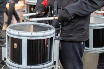 A young man wearing dark clothing holds two drumsticks and beats on a black and white drum. There are multiple drummers in the area.