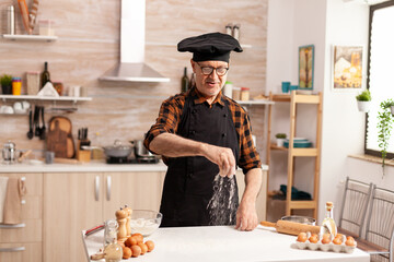 Senior baker preparing bread using bio wheat flour in home kitchen wearing apron and bonete. Cook in kitchen uniform sprinkling sieving sifting ingredients by hand.