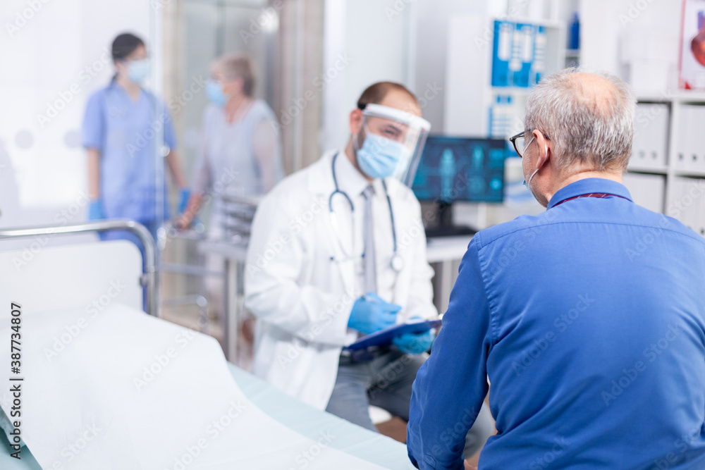 Old patient listening doctor diagnosis while wearing protective mask against coronavirus outbreak in hospital examination room. Modern private clinic . Practitioner physician appointment. - obrazy, fototapety, plakaty 