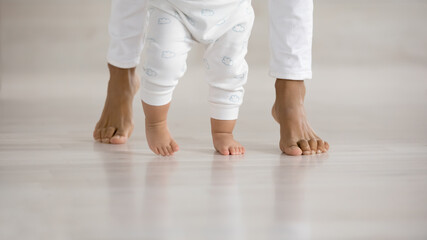 Wall Mural - crop close up of little baby infant learn walking at home wooden floor holding mom hands. cute small