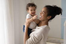 Smiling Young African American Mom Hold In Hands Happy Little Ethnic Baby Infant Play At Home Together. Happy Biracial Mother Hug Embrace Feel Playful With Small Toddler Kid Child Show Love Care.