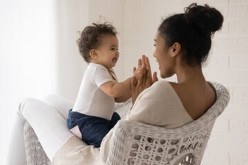 Wall Mural - Smiling young biracial mom sit in chair at home have fun engaged in funny game with little toddler baby. Happy african American mother relax play with small newborn infant child. Childcare concept.