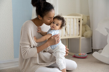 Wall Mural - Happy african American young mother kiss cute little baby infant show love and care at home. Smiling biracial mom hug cuddle play with small toddler child in children room. Motherhood concept.