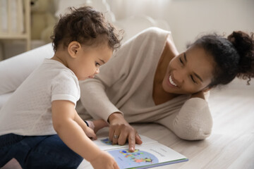 Wall Mural - smiling loving african american mom lying on warm floor in children room reading book with curious b