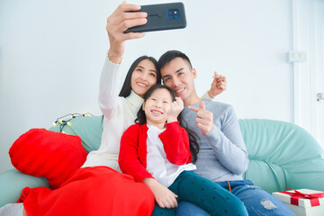 Wall Mural - Beautiful asian mom, dad and daughter are doing selfie via smart phone and smiling while celebrating chrstmas holiday.