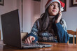 © AliciaFdez - Una chica joven habla por su teléfono móvil mientras lleva un gorro de Santa Claus y un jersey de Navidad frente a su ordenador portátil