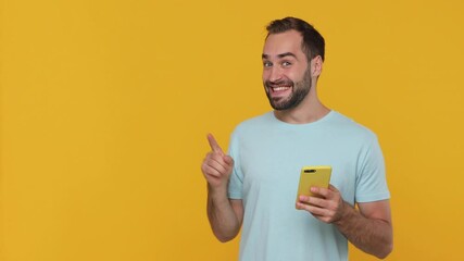 Canvas Print - Shocked surprised young man in basic casual blue t-shirt say wow hold in hand using mobile cell phone pointing fingers aside on workspace isolated on yellow background studio. People lifestyle concept