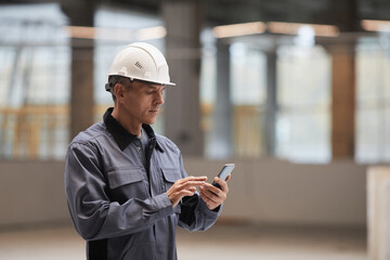 side view portrait of mature worker using smartphone while standing at construction site or in indus