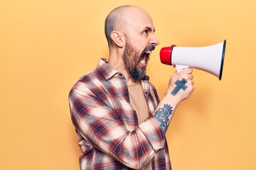 Young handsome bald man with angry expression. Screaming loud using megaphone standing over isolated yellow background