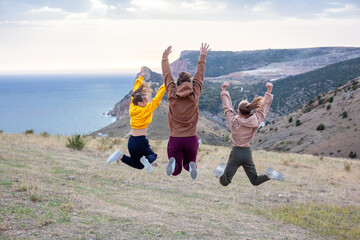 Back view of two preteen girls and young woman traveler wearing yellow and biege hoodie jumping on top of the mountain landscape and looking to the sea