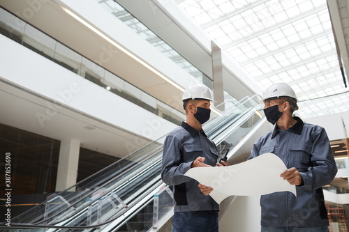 Low angle portrait of two construction workers wearing masks and discussing plans while standing in shopping mall or office building, copy space