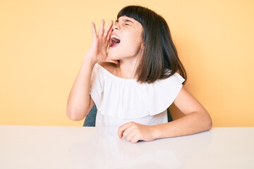 Wall Mural - Young little girl with bang wearing casual clothes sitting on the table shouting and screaming loud to side with hand on mouth. communication concept.