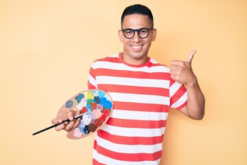 Wall Mural - Young handsome latin man holding paintbrush and palette smiling happy and positive, thumb up doing excellent and approval sign