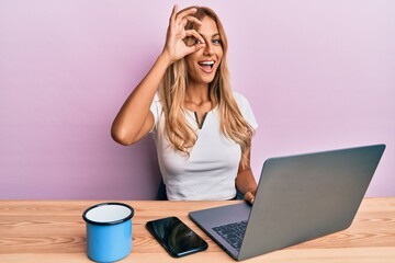 Sticker - Beautiful blonde young woman working using computer laptop smiling happy doing ok sign with hand on eye looking through fingers