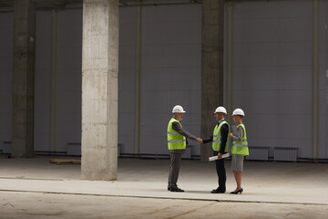  Wide angle portrait of business people shaking hands while discussing investment deal at construction site indoors, copy space