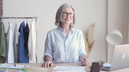 Wall Mural - Happy middle aged stylish woman fashion designer wears glasses standing at workplace desk, portrait. Smiling sophisticated 60s old lady dressmaker drawing sketches, looking at camera in modern atelier