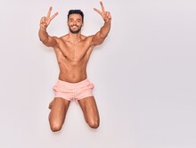 Young Handsome Hispanic Man On Vacation Wearing Swimwear Shirtless Smiling Happy. Jumping With Smile On Face Doing Victory Sign Over Isolated White Background