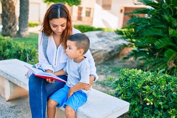 Wall Mural - Adorable latin mother and son sitting on the bench and reading book at the park.