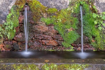 Long exposure of water coming out of a fountain in the wall, covered in moss