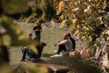 Shallow Focus On The Branches In Foreground, Outline Of A Group Of People In The Background, Sitting On The Grass Outside, Travelling In Nature In Autumn. Friends Gathering For A Barbecue In The Widl