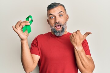 Wall Mural - Middle age handsome man holding support green ribbon pointing thumb up to the side smiling happy with open mouth