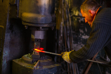 A blacksmith forges an iron rod with a mechanical hammer. Work in the forge