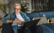 Focused Elderly Man Writing Notes In Notebook Watching Webinar Or Online Training Using Laptop Computer, Modern Senior Male With Gray Hair And Beard Learning Online