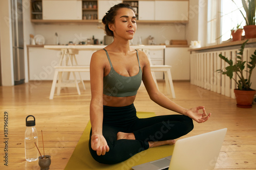 Fit young Latin woman meditating before yoga to calm her mind, keeping eyes closed, making mudra gesture, sitting in front of open laptop in half lotus pose, listening to video instructor online