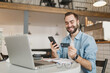 © ViDi Studio - Smiling young man sit alone at table in coffee shop cafe restaurant indoor working or studying on laptop pc computer hold credit bank card using cell phone. Freelance mobile office business concept.