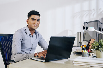 Wall Mural - portrait of smiling indian programmer working on laptop at office desk
