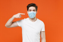 Young Man 20s Wearing Casual White T-shirt Pointing Index Finger On Sterile Face Mask To Safe From Coronavirus Virus Covid-19 During Pandemic Quarantine Isolated On Orange Background Studio Portrait.