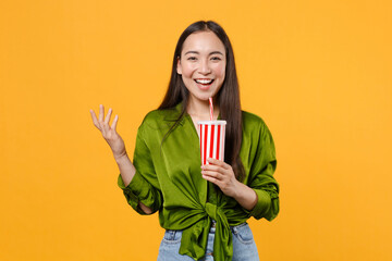 Wall Mural - Funny young brunette asian woman wearing basic green shirt standing holding plastic cup of cola or soda spreading hands looking camera isolated on bright yellow colour background, studio portrait.