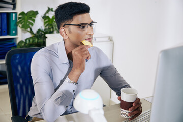 Wall Mural - pensive young indian software developer in glasses drinking coffee, eating cookie and checking progr