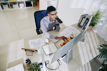 Wall Mural - indian programmer in glasses checking code on computer screen and recording voice message for cowork