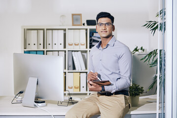 Wall Mural - portrait of confident young indian entrepreneur sitting on edge of office desk with tablet computer 