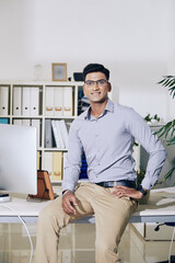 Wall Mural - handsome young indian entrepreneur sitting on edge of office desk and smiling at camera