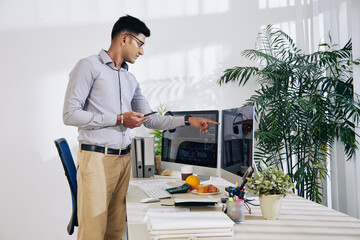 Wall Mural - pensive indian programmer with smartphone in hand pointing at computer screen with programming code 
