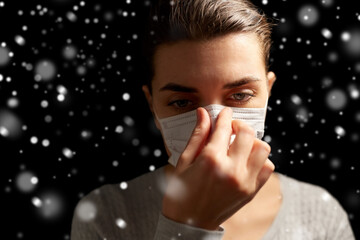 Wall Mural - health protection, safety and pandemic concept - sick young woman adjusting protective medical face mask over snow on black background in winter