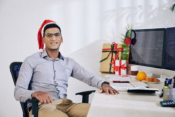 Wall Mural - portrait of positive indian software developer in santa claus hat sitting at office desk