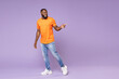 © ViDi Studio - Full length of smiling cheerful young african american man 20s wearing basic casual orange t-shirt standing pointing index finger aside isolated on pastel violet colour background, studio portrait.