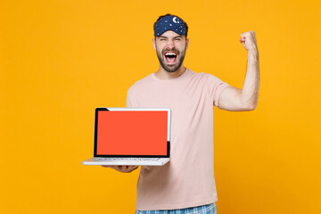 Happy young man in pajamas home wear sleep mask hold laptop pc computer with blank empty screen doing winner gesture resting at home isolated on yellow background. Relax good mood lifestyle concept.