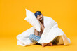 © ViDi Studio - Full length of smiling young bearded man in pajamas home wear sleep mask sitting with pillow blanket isolated on bright yellow colour background studio portrait. Relax good mood lifestyle concept.