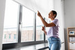 © Syda Productions - people and leisure concept - happy young african american woman opening window roller blinds at home