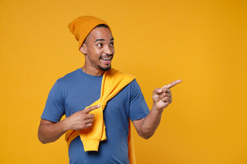 Wall Mural - Smiling cheerful funny young african american man 20s in basic casual blue t-shirt hat pointing index fingers aside on mock up copy space isolated on bright yellow colour background, studio portrait.