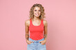 Smiling charming beautiful attractive young blonde woman 20s wearing basic casual tank top standing holding hands in pockets looking camera isolated on pastel pink colour background, studio portrait.