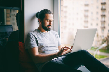 Handsome attractive young dark-haired man working on laptop at home while sitting on windowsill with city view